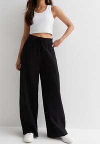 Zwarte wijde broek van textiel met een elastische tailleband en een trekkoord. Gecombineerd met een witte cropped tanktop en witte sneakers.