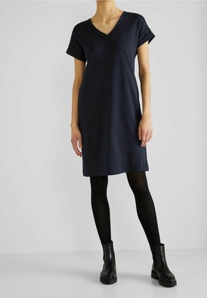 Vrouw die een marineblauwe, korte mouw V-hals jurk draagt met zwarte panty's en zwarte enkellaarzen, staand tegen een effen achtergrond.