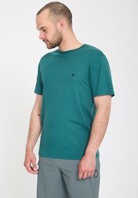 Homme aux cheveux courts et barbe, portant une chemise à manches courtes bleu sarcelle et un short gris, debout et regardant vers le bas à sa gauche.