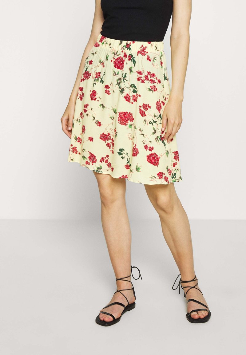 Květinová sukně se žlutým základem, zdobená červenými a zelenými vzory květin. Elastický pas a rozšířený střih. Doplňena černými sandály na tenkých páscích.