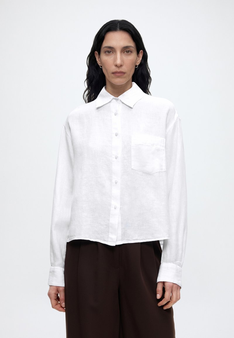 Femme aux longs cheveux noirs portant une chemise blanche boutonnée avec une poche poitrine et un pantalon large marron foncé, debout devant un fond blanc.