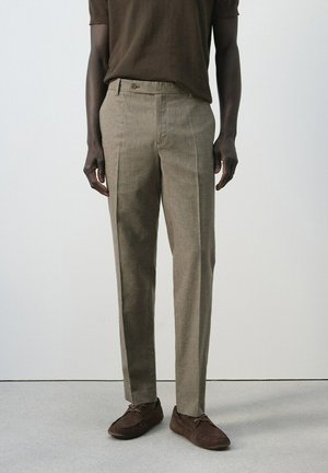 Mężczyzna ubrany w beżowe spodnie na miarę, ciemnobrązowe zamszowe buty oraz ciemnobrązową koszulę z krótkim rękawem, stojący na tle gładkiej jasnej ściany.