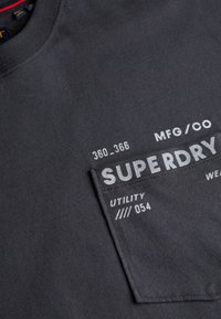 Dunkelgraue Hemdtasche mit weißem, aufgedrucktem Text, der „SUPERDRY“, „MFG/CO“, „360_366“ und „UTILITY //// 054“ auf dem Stoff zeigt.