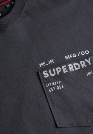 T-shirt in cotone grigio scuro con collo rotondo, taschino sul petto e testo stampato bianco. Il design include loghi e dettagli funzionali.