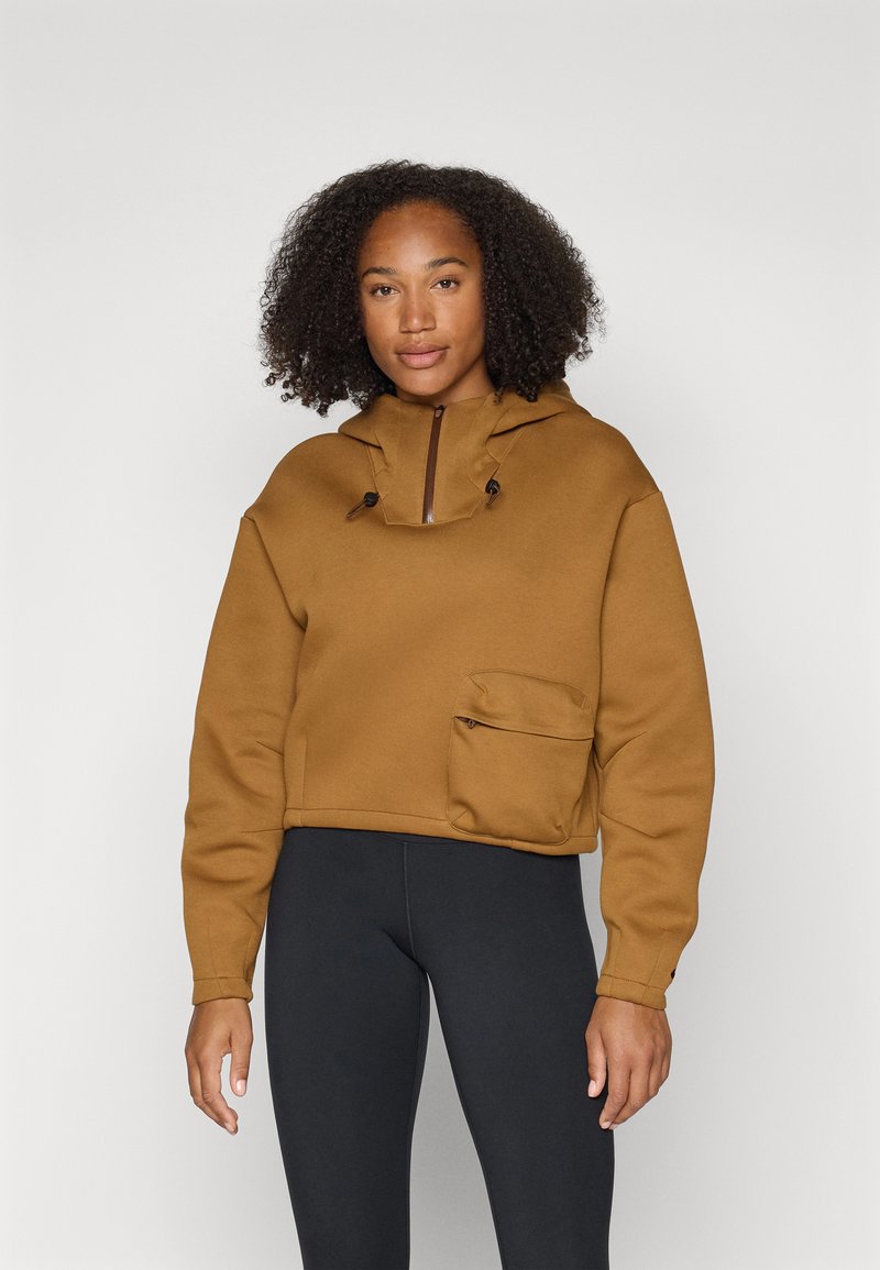 Brauner Kapuzenpullover aus weichem Stoff, mit einem halben Reißverschluss, verstellbaren Kordeln und einer Fronttasche. Kombiniert mit schwarzen Leggings.