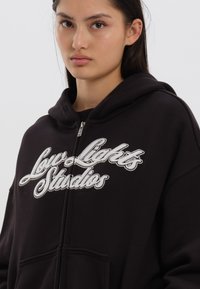 Sweatshirt preto com fecho éclair, feito de algodão, apresentando um logótipo bordado em branco "Low Lights Studios" na frente. Inclui um bolso frontal espaçoso.