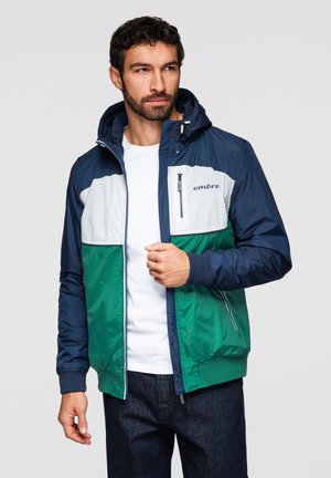 Homme aux cheveux foncés et à la barbe portant une veste à capuche bleu marine, blanche et verte par-dessus une chemise blanche et un jean foncé, regardant sur le côté.