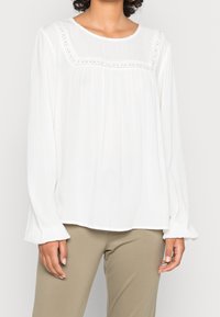 Blusa bianca a maniche lunghe con tessuto strutturato, arricchita da delicati dettagli in pizzo lungo la scollatura e maniche arricciate. Abbinata a pantaloni color kaki.
