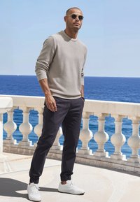 Hellgrauer Strickpullover, dunkle Hose mit elastischen Bündchen und weiße Sneaker. Sonnenbrille vervollständigt das Outfit vor einer Küstenkulisse.