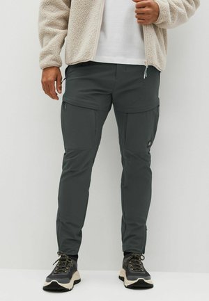 Mężczyzna ubrany w ciemnoszare spodnie bojówki, czarno-białe trampki, białą koszulę oraz beżową kurtkę z polaru, stojący na tle jednolitego jasnego tła.