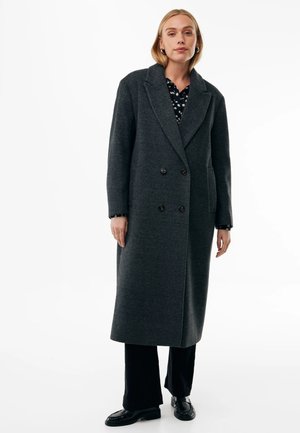Donna che indossa un lungo cappotto doppiopetto grigio scuro sopra pantaloni neri e scarpe nere, in piedi con un'espressione neutra su uno sfondo bianco.