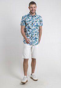 Chemise à manches courtes avec un motif de feuilles bleu et vert, associée à un short blanc. Le modèle porte des baskets blanches avec des accents beige.