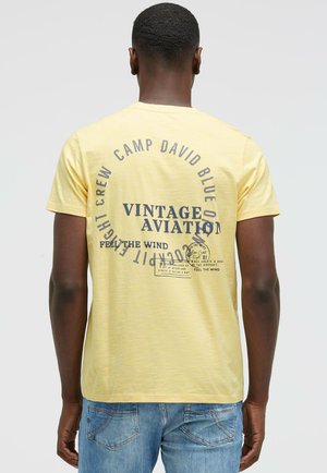 Gelbes Kurzarm-T-Shirt mit einer Vintage-Flugzeuggrafik und dunkelblauem Text auf dem Rücken, aus weichem Baumwollstoff gefertigt.
