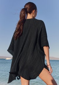 Črni kaftan s črtami, ohlapnega kroja, kratkimi rokavi in poševnim robom. Teksturiran material z raztrganimi robovi, nošen na plaži.