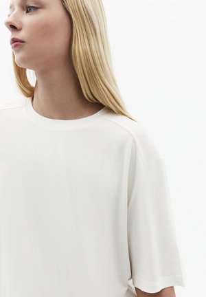 Junge Frau mit langen blonden Haaren, die ein einfaches weißes, übergroßes kurzärmeliges Shirt trägt und nach links schaut, vor einem weißen Hintergrund.