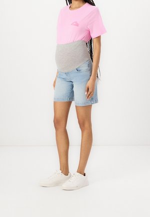 Femme enceinte portant un t-shirt rose, une ceinture de maternité grise, un short en jean bleu clair et des baskets blanches, debout devant un fond blanc.