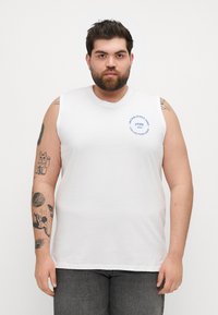 T-shirt sans manches blanche en coton, affichant un logo graphique bleu sur la poitrine gauche. Design simple et décontracté avec un col rond.