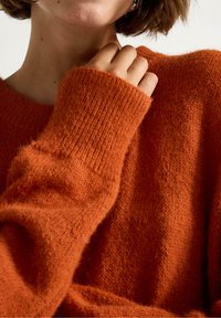 Donna che indossa un morbido maglione arancione oversized con maniche lunghe, mano che tocca delicatamente il colletto vicino al collo su uno sfondo neutro.