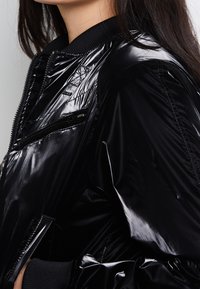 Veste bomber noire en matériau brillant avec une poche zippée, un col côtelé et des détails texturés sur les épaules. Design léger et ajusté.