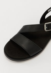 Sandálias de couro preto com tiras cruzadas, palmilha texturizada preta e detalhe em fivela de metal prateado. Sola plana com um design minimalista.