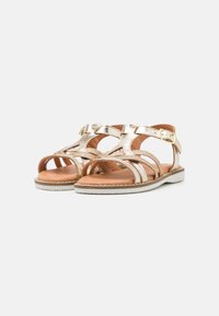 Sandali metallici oro con cinturini incrociati, design open toe, fibbia regolabile alla caviglia e soletta tan imbottita su una suola esterna bianca.