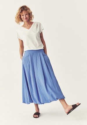 Kihara juustega naine, kes kannab valget lühikese varrukaga topi, sinist midi seelikut ja musti libisevaid sandaale, seisab ühe jalaga tõstetud.