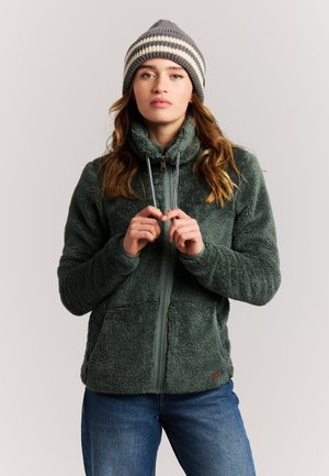 Frau trägt grüne flauschige Reißverschlussjacke und gestreifte Strickmütze, hält Jackenzüge, steht vor einfarbigem Hintergrund.