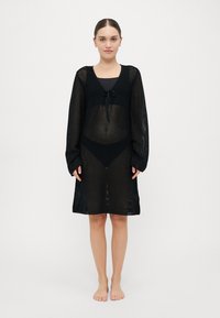 Black sheer knit cover-up dress with long sleeves, featuring a tie at the neckline and a loose fit, over a black bikini bottom.