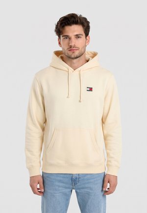 Ung mand iført en lysebeige hættetrøje med frontlomme og Tommy Jeans-logo, kombineret med lyseblå jeans, står mod en ensfarvet baggrund.
