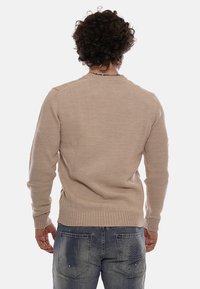 Uomo con capelli ricci che indossa un maglione beige e jeans blu con il logo DS2, in piedi rivolto di spalle contro uno sfondo neutro.