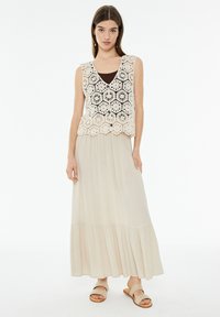 Top de encaje de ganchillo en color crema sobre una camiseta negra, combinado con una falda larga y fluida en crema con dobladillo en capas y sandalias beige.
