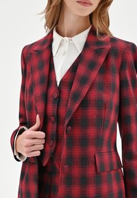 Completo a quadri rosso e nero con blazer aderente, doppio bottone e gilet abbinato; abbinato a una camicia bianca con colletto sotto.