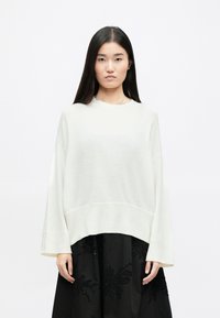 Light cream, oversized sweater with a ribbed hem and cuffs. Soft texture, rounded neckline, and dropped shoulders. Worn over a black skirt.