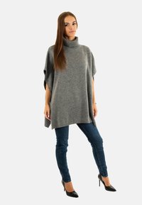 Poncho de punto gris con cuello alto, con un corte holgado y aberturas laterales. Combinado con jeans ajustados azul oscuro y tacones altos negros.