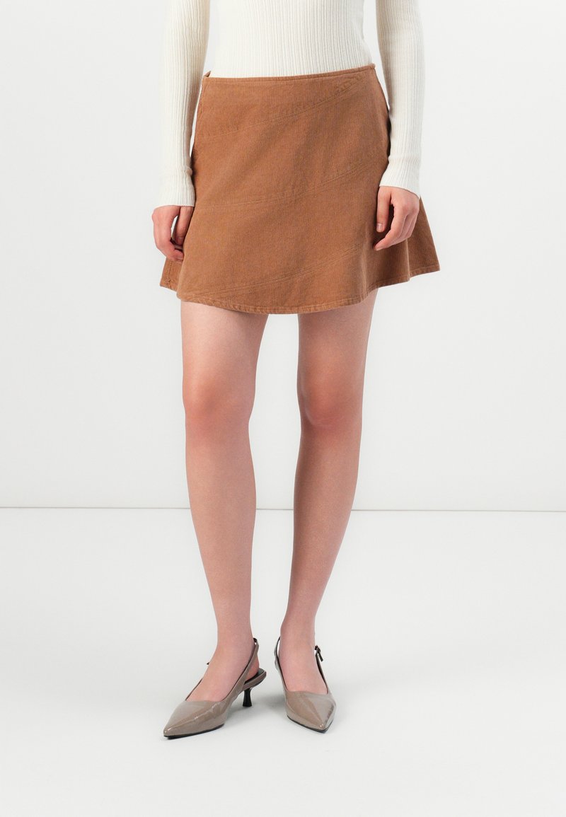 Donna che indossa una mini gonna in velluto a coste marrone, una maglietta bianca a maniche lunghe a costine e delle scarpe slingback beige con tacco kitten, in piedi su un pavimento bianco.