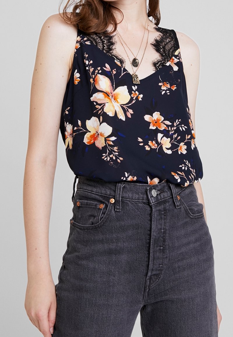 Blommig ärmlös topp i marinblått med orange och vita blommor, med en spetskantad halsringning. Matchad med höga mörkgrå jeans.