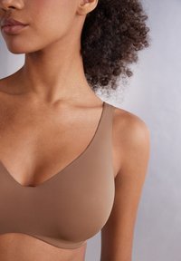 Hnědá bezšvová podprsenka vyrobená z hladké tkaniny, s širokými ramínky a měkkým designem ve tvaru písmene V, na neutrálním pozadí.