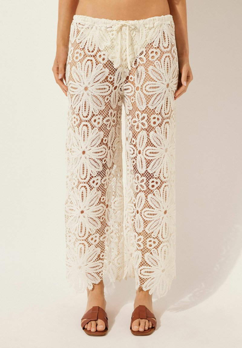 Pantalones palazzo de encaje en color crema con estampado floral, cintura con cordón y textura translúcida, combinados con sandalias planas marrones.