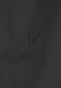 Černá tkanina s jemným vyšitým logem "K" ve stejném odstínu, která se vyznačuje hladkou texturou a jednoduchým designem.