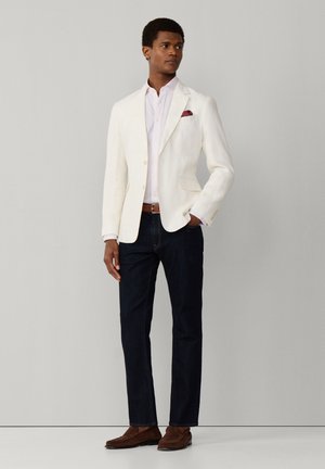 Man die een witte blazer, lichtroze overhemd, donkere jeans, een bruine riem en bruine loafers draagt, staand tegen een effen grijze achtergrond.
