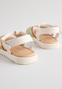 Sandales blanches avec accents rayés colorés, sangles réglables et semelles texturées. L'intérieur présente une semelle beige avec des coutures décoratives.