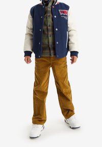 Námořnická a krémová varsity bunda přes zelenou kostkovanou košili; béžové kordové kalhoty; bílé tenisky; obsahuje knoflíky a logo detaily.