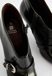 Musta lakattu nilkkaboots, jossa on sivuvetoketju, kapea korko ja koristetukka. Sisävuori burgundin värinen, koko 40. Logot näkyvät sisällä.