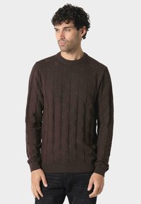 Brauner Pullover mit einem geometrischen Rautenmuster. Runder Ausschnitt, lange Ärmel und gerippte Bündchen. Der Stoff wirkt strukturiert und gestrickt.