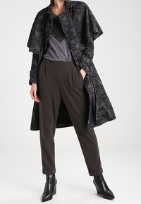 Černý a šedý texturovaný kabát s oversized rukávy, přetahovaný přes tmavě šedé tričko s výstřihem do V a tmavé kalhoty. Černé kotníkové boty.