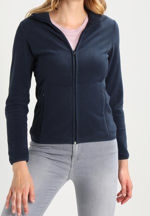 Frau trägt eine enganliegende marineblaue Fleecejacke mit Reißverschluss über einem hellrosa Hemd und hellgrauen Jeans, steht mit entspannt an den Seiten hängenden Händen.