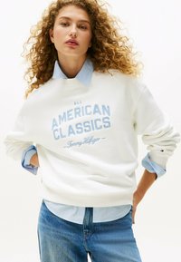Witte sweatshirt met blauwe geborduurde tekst "ALL AMERICAN CLASSICS" en merknaam. Draagover een lichtblauw overhemd met kraag. Spijkerbroek zichtbaar.
