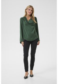 Blouse en satin vert foncé avec un col drapé à effet de cowl et des manches longues, associée à un jean skinny noir et des mules noires à bout pointu.