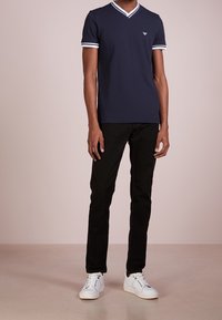 Navy modrá V-krční tričko s bílými pruhy na rukávech, v kombinaci s černými kalhotami a bílými teniskami. Hladký bavlněný materiál.