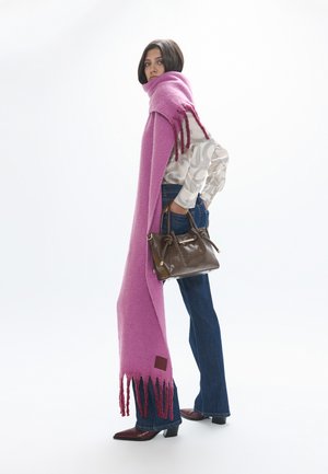 Mujer con una larga bufanda rosa con flecos, blusa blanca estampada, vaqueros azules, bolso marrón y botines, de pie frente a un fondo blanco.
